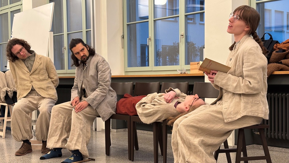 Schauspielende proben eine Szene, drei Figuren Sitzen auf einem Stuhl, eine liegt zwischen ihnen auf mehreren.