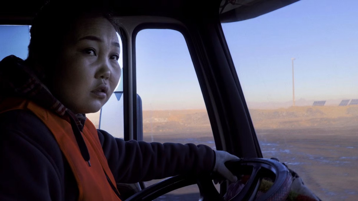 Eine mongolische Frau sitzt in einem LkW am Steuer. Sie fährt durch eine karge Landschaft.